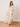 A woman wearing a long-sleeve maxi dress with a colorful geometric checkered pattern in pink, orange, yellow, blue, and green on a white base, standing on a wooden floor against a plain off-white wall.