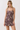 A woman modeling a sleeveless mini dress with a brown and beige botanical print and pink button accents, photographed against a plain light gray studio background.