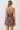 A woman viewed from the back wearing a brown mini dress with a botanical and bird pattern, featuring thin crisscross straps and a flared skirt, set against a plain light background.