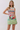 A woman modeling a sleeveless mini dress with a colorful, painterly art print depicting a crowd scene and a boxer, set against a plain light gray studio background.