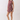 A woman modeling a sleeveless ruffled romper in a deep burgundy color with a beige and green floral pattern, paired with beige block-heel sandals, against a plain light gray studio background.