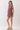 A woman modeling a sleeveless ruffled romper in a deep burgundy color with a beige and green floral pattern, paired with beige block-heel sandals, against a plain light gray studio background.
