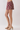 A person wearing patterned shorts in a burgundy base with gold, green, and beige paisley motifs, paired with beige heeled sandals, shown from the waist down against a light gray studio background.