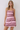 A woman modeling a sleeveless, scoop-neck mini dress with horizontal pink, magenta, and burgundy stripes against a plain light gray studio background.