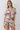 A woman modeling a short-sleeve floral romper with a tie-front neckline and ruffled edges, photographed against a plain light gray studio background.