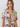 A woman wearing a short-sleeve tie-front blouse with a colorful tropical floral print on a light cream background, photographed against a plain light gray studio backdrop.