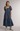 A woman modeling a tiered midi dress with a blue, green, and purple plaid pattern, paired with beige ankle boots, against a neutral studio background.