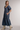 A woman modeling a long plaid midi dress with puff sleeves, a scoop neckline, and tiered skirt, paired with beige ankle boots against a neutral studio background.