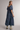 A woman modeling a tiered puff-sleeve midi dress with a blue, purple, and green plaid pattern, paired with light beige cowboy boots, against a neutral gray studio background.