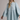 A woman wearing a light blue denim shirt dress with long bell sleeves, a buttoned collar, and a gathered waist, photographed against a plain light gray studio background.