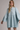 A woman wearing a light blue denim shirt dress with long bell sleeves, a buttoned collar, and a gathered waist, photographed against a plain light gray studio background.