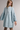 A woman wearing a light blue denim-style mini dress with a pointed collar, button placket, gathered waist seam, and dramatic bell sleeves, photographed against a plain light gray studio background.
