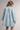 A woman shown from the back wearing a light blue denim-style mini dress with long bell sleeves, gathered seams, and a collared neckline, photographed against a plain light gray studio background.