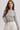 A woman wearing a gray and white horizontally striped long-sleeve knit top with white drawstring pants, photographed against a light gray studio background.