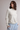 A woman wearing a cream and light blue striped long-sleeve mock neck top and light blue textured pants, standing against a neutral light gray studio background.