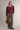 A woman stands against a light gray studio background wearing a burgundy and white horizontally striped long-sleeve knit polo sweater, brown wide-leg pants, olive green boots, and holding a brown leather handbag.