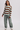 A woman modeling a knit sweater with horizontal brown and pale mint green stripes, paired with wide-leg sage green pants and white sneakers, against a light gray studio background.