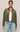 A woman modeling a cropped, long-sleeve button-front overshirt in olive green suede-like fabric, worn over a white tank top and light-wash blue jeans against a plain light beige studio background.