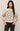 A woman modeling a cream-colored knit sweater with horizontal black and gray stripes, paired with black jeans, against a plain light beige studio background.