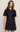A woman modeling a dark indigo denim mini shirt dress with short sleeves, gold-tone buttons, and front patch pockets with decorative gold hardware, set against a plain light beige studio background.