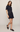 A woman models a dark indigo denim mini dress with short sleeves, a button-up front, and two front patch pockets with button closures. She is wearing dark brown leather loafers against a plain, light beige studio background.
