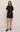 A woman modeling a dark indigo denim mini dress with short sleeves, a button-up front, and two front patch pockets with gold-tone hardware, paired with dark brown leather loafers, against a plain light beige studio background.