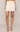 A model wearing a cream-colored mini skirt with floral lace trim along the hem, paired with nude high-heeled sandals against a light beige studio background.