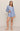 A woman modeling a light blue long-sleeve pajama set with a small dark floral print, featuring a cropped tie-front top and matching shorts, against a plain beige studio background.