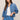 A woman modeling a cropped blue denim jacket over a white tank top and matching blue denim drawstring pants, photographed against a plain beige studio background.