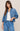 A woman modeling a cropped blue denim jacket over a white tank top and matching blue denim drawstring pants, photographed against a plain beige studio background.