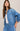 A woman wearing an oversized light blue denim jacket over a white ribbed top and matching blue denim bottoms, photographed against a plain warm beige studio background.