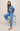 A woman models a matching blue denim set consisting of a cropped button-up shirt and wide-leg cropped pants with white side stripes, seated on a metal stool against a neutral beige background.