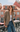 A woman stands on a balcony wearing an oversized brown and beige houndstooth blazer over a white tank top and blue jeans, with a pink building in the background.