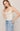 A woman modeling a light beige ribbed camisole tank top with delicate lace trim along the scoop neckline and shoulder straps, paired with light-wash blue jeans against a solid light beige background.