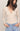 A woman wearing a beige long-sleeve V-neck top with a small cutout at the center chest and light blue jeans, photographed against a plain light background.