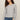 A woman modeling a light gray long-sleeve knit top with a wide boat neckline, paired with blue jeans, against a plain beige studio background.