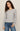 A woman modeling a light gray long-sleeve knit top with a wide boat neckline, paired with blue jeans, against a plain beige studio background.