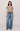 A model stands against a light gray studio background wearing a taupe short-sleeve knit top, blue wide-leg jeans, a brown belt, and dark brown shoes.