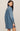 A woman modeling an oversized blue denim shirt dress, shown from a side-back angle against a plain beige studio background.