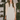 A woman models a short white satin slip dress with a lace hem, styled with a cream-colored knit cardigan. She is standing in a boutique setting with shelves of shoes and handbags and a plant in the background.