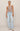 A model stands against a plain beige studio background wearing a sleeveless taupe knit top, light-wash wide-leg jeans, and white sneakers.