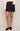 A dark wash denim mini skirt with gold-tone hardware and contrast stitching, worn by a model in a cream knit sweater against a light gray studio background.