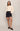 A model stands against a light gray studio background wearing a cream-colored, textured knit sweater, a dark blue denim mini skirt, and black leather loafers.
