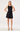 A blonde model wearing a sleeveless black satin-like mini dress with a draped neckline and a flared skirt, paired with gold strappy heels, against a plain white studio background.