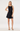 A woman modeling a sleeveless black mini dress with a draped neckline and a flared skirt, paired with gold strappy high heels, against a plain light gray studio background.