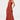 A woman modeling a sleeveless, high-neck, floor-length gown in a warm rust or terracotta color, photographed against a clean light gray studio background.