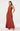A woman modeling a sleeveless, high-neck, floor-length gown in a warm rust or terracotta color, photographed against a clean light gray studio background.