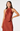 A woman modeling a sleeveless, high-neck satin dress in a warm rust or terracotta color, photographed against a clean light gray studio background.