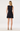 A woman viewed from the back wearing a sleeveless black mini dress with a gathered waist and flared skirt, paired with gold high-heeled sandals, against a plain light gray studio background.