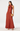 A woman models a sleeveless rust-orange satin halter maxi dress with a structured waist, holding a textured gold clutch against a plain white studio background.
